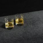 Two glasses of whiskey on a gray surface in front of a black wall.